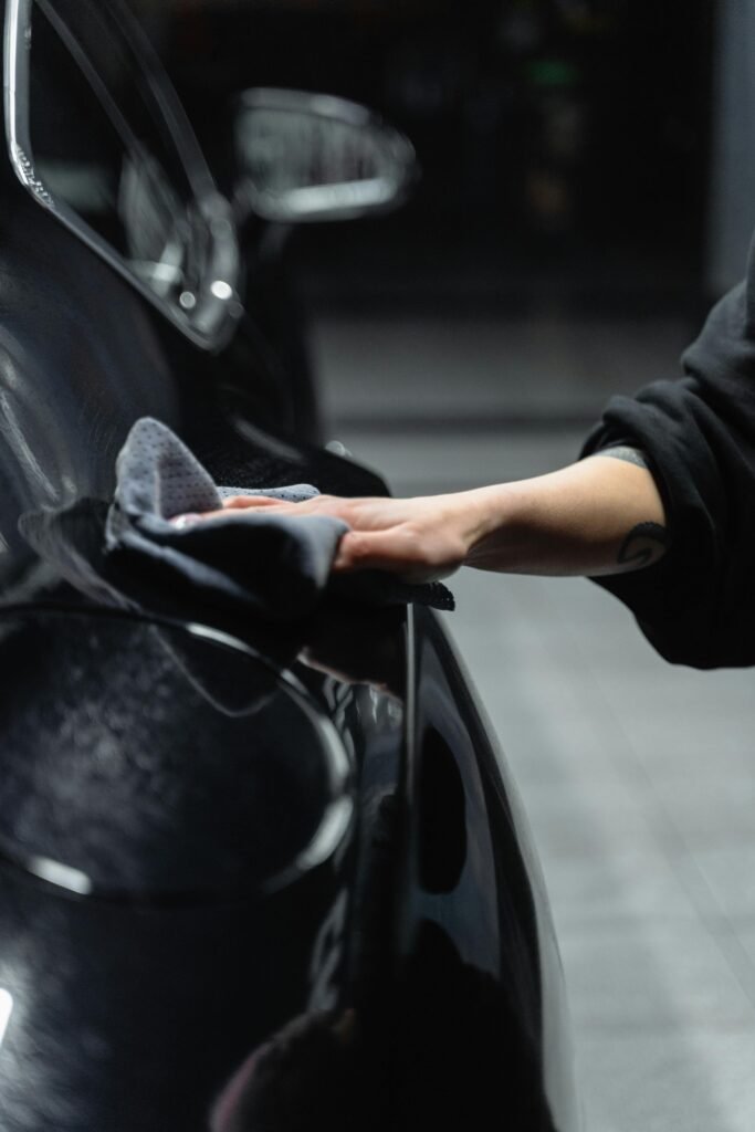 Close-up of a person's hand cleaning a shiny black car with a microfiber cloth in a garage.